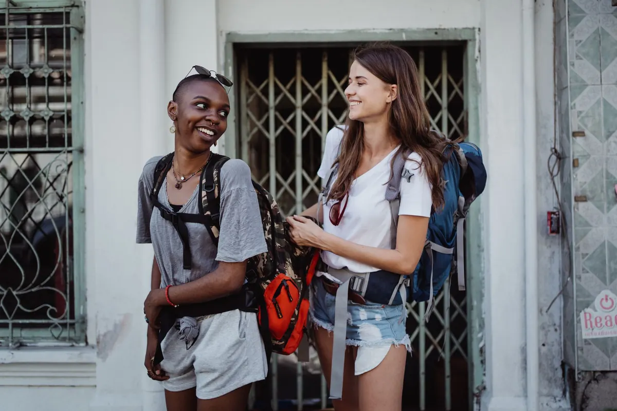 Two women with backpacks laughing together on a city street, one adjusting the other's pack