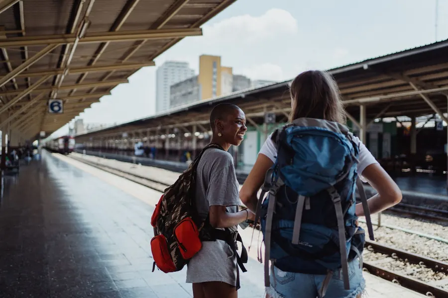 Two women with backpacks talking on a railway platform, one in profile and one seen from behind